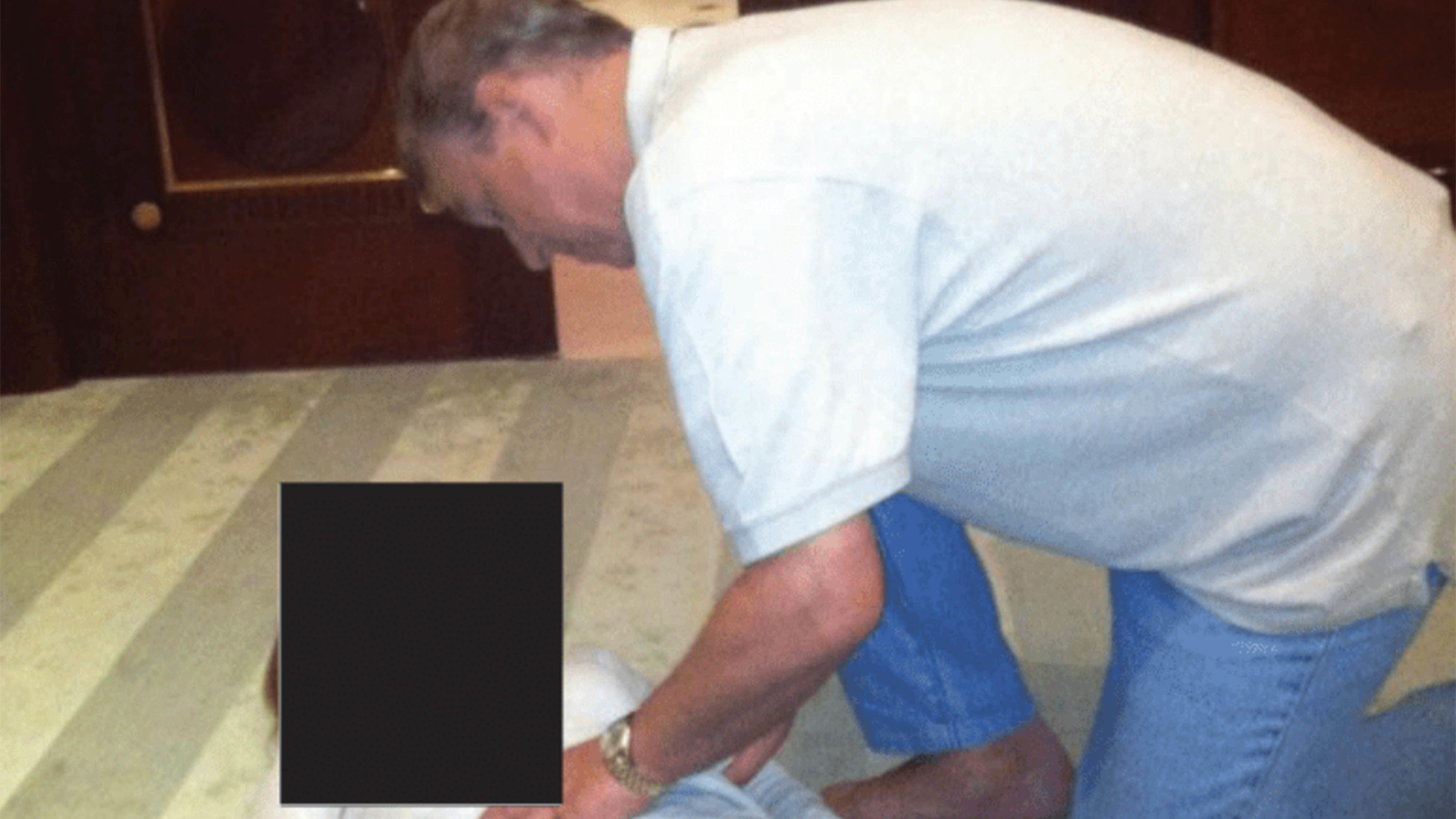 Um homem com uma camisa polo branca e cal&ccedil;a jeans azul desbotada est&aacute; de p&eacute; sobre uma mulher com cal&ccedil;a preta e blusa branca. O rosto dela est&aacute; oculto por um ret&acirc;ngulo preto. O homem est&aacute; tocando a barriga dela. Ela est&aacute; deitada no ch&atilde;o de uma sala grande, provavelmente uma sala de jantar ou um corredor.