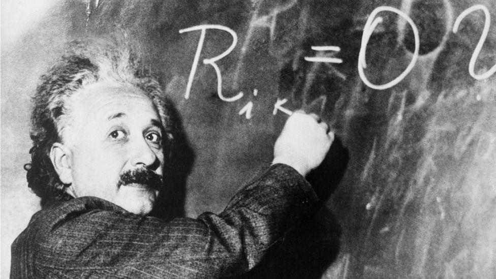 Albert Einstein mirando la cámara mientras escribe una fórmula matemática en un pizarrón.