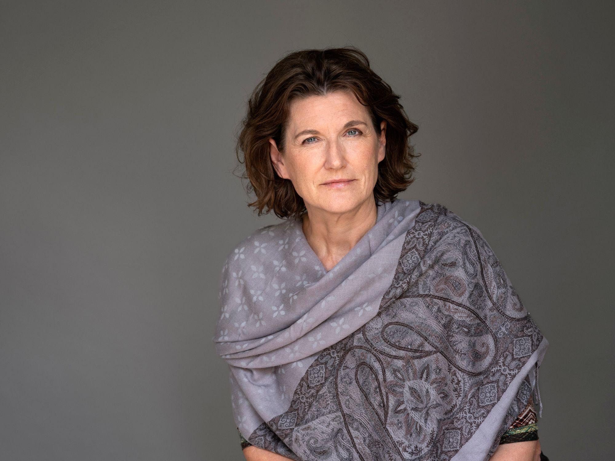Mujer de cabello corto y ondulado vestida con un chal gris de estampado floral y paisley, posando frente a un fondo neutro.