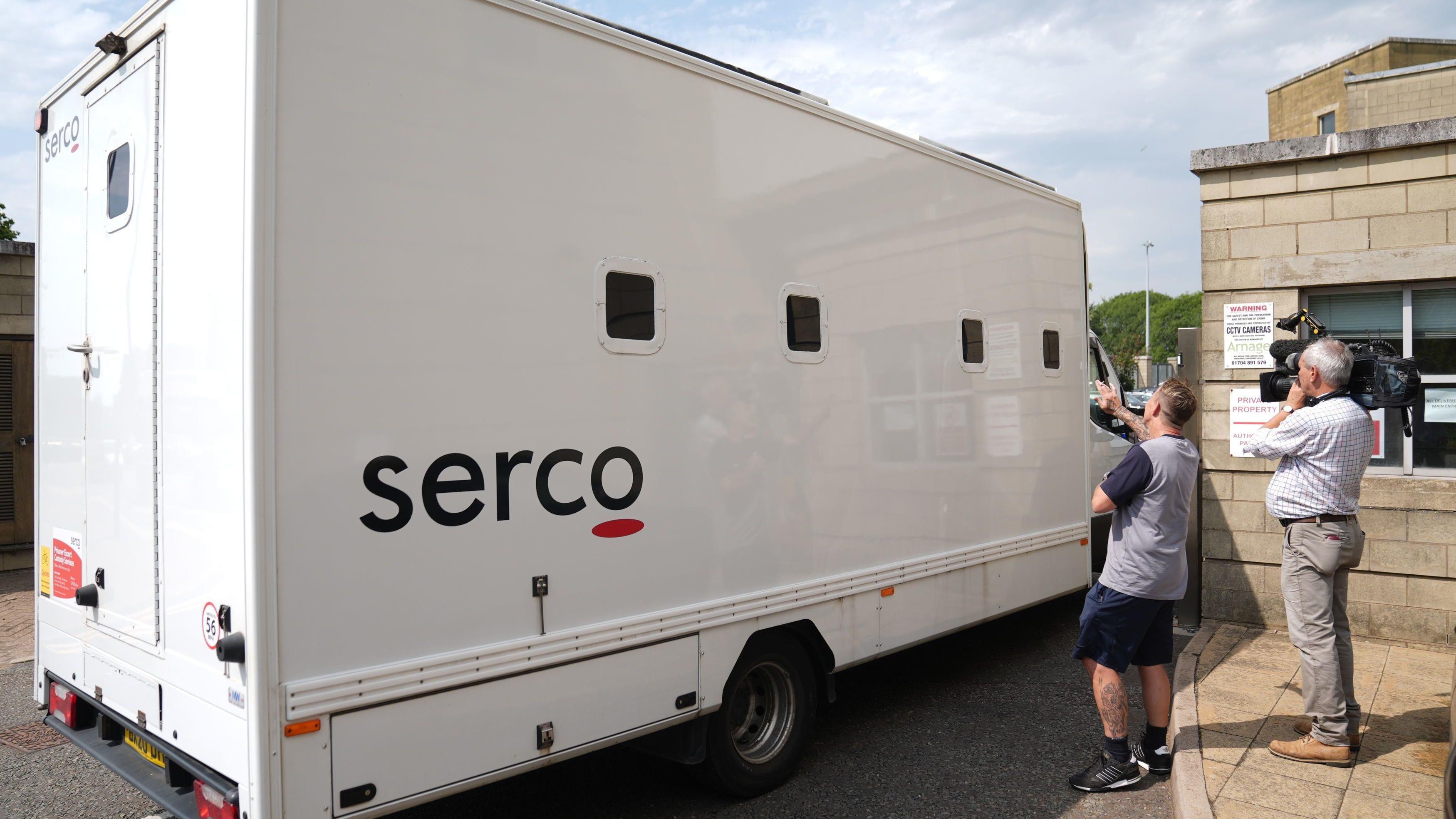 شاحنة سجن بيضاء بأربع نوافذ صغيرة، مكتوب عليها اسم "سيركو" على جانبها، تمر عبر بوابات محكمة. ويظهر أيضاً في الصورة رجلان، أحدهما يرتدي شورتاً وقميصاً، ويبدو أنه يلوّح للشاحنة بينما يقوم الآخر وهو مصور أخبار تلفزيونية، بتصوير الشاحنة وهي متجهة إلى المحكمة. 