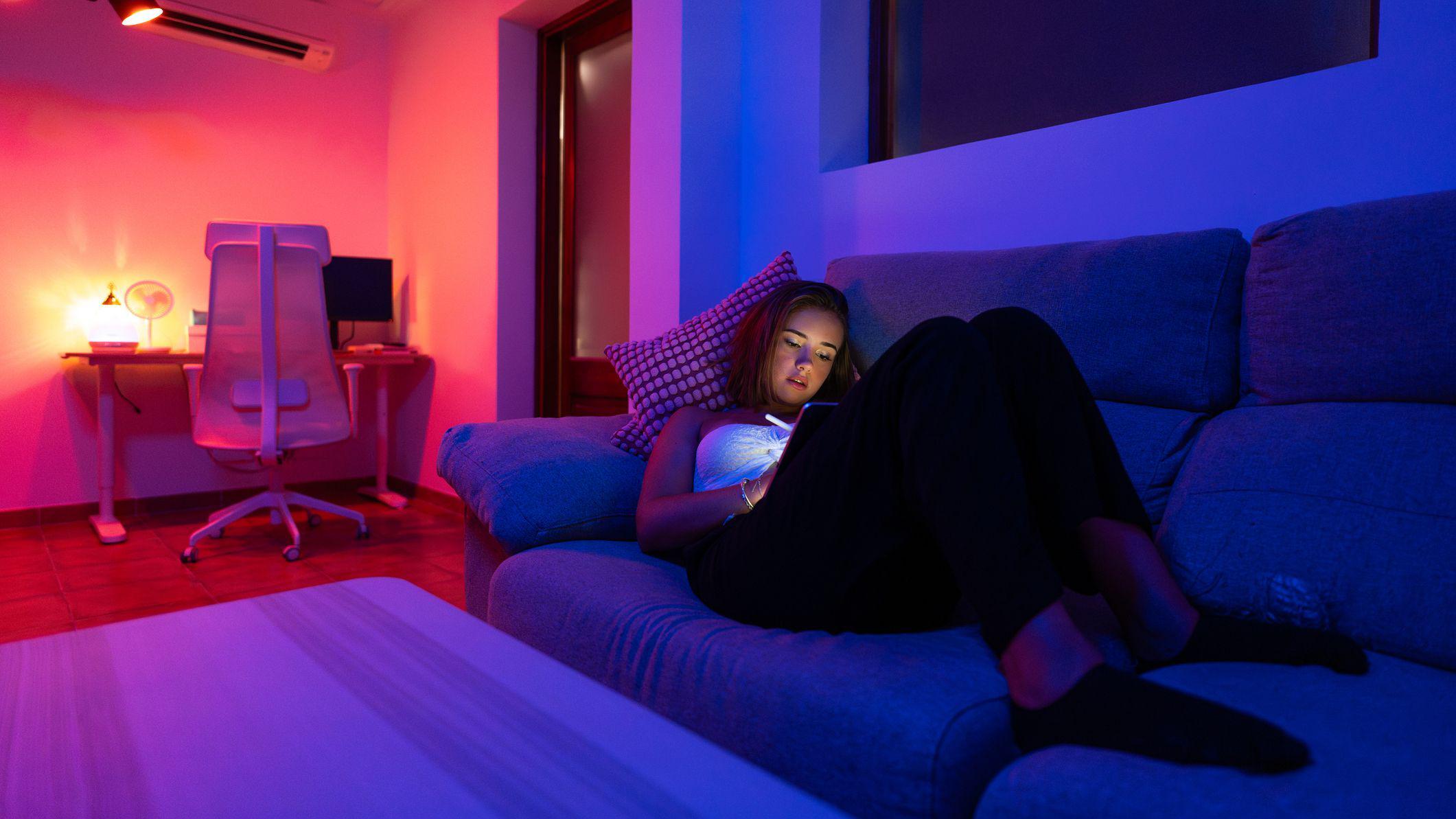 Jovem recostada no sof&aacute; olhando para uma tela, com as luzes da sala reduzidas e uma mesa com abajur aceso ao fundo