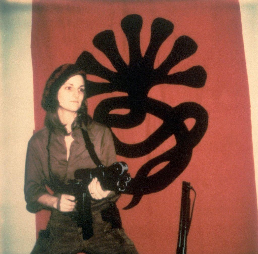 Patty Hearst, vestida con camuflados, tirantas y una boina negra, sostiene un arma semiautomática, frente al escudo del SLA: una cobra negra de siete cabezas.