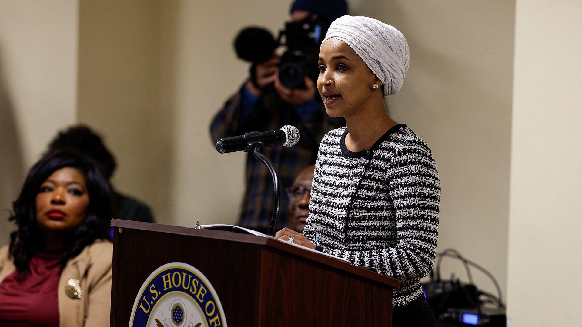 Una fotografía de Ilhan Omar dirigiéndose a una reunión municipal  vestida con jersey de rayas negras y blancas, turbante blanco. Habla por un micrófono en un podio con otras personas en el fondo; el podio muestra un logotipo.