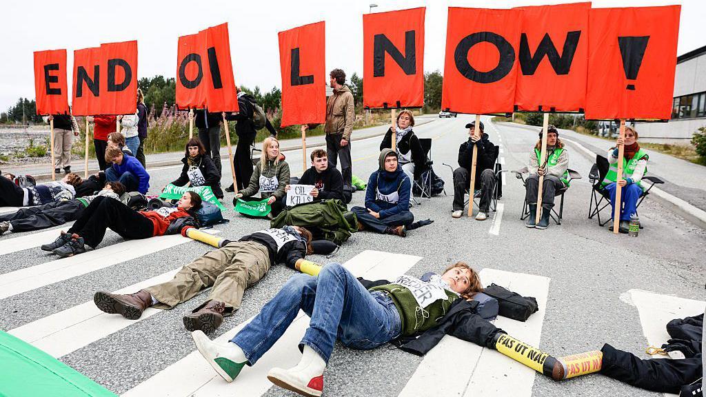 Um grupo de jovens protesta contra a explora&ccedil;&atilde;o petrol&iacute;fera na Noruega. Alguns est&atilde;o deitados no asfalto, outros sentados em cadeiras segurando cartazes com letras que, unidas, formam a mensagem 