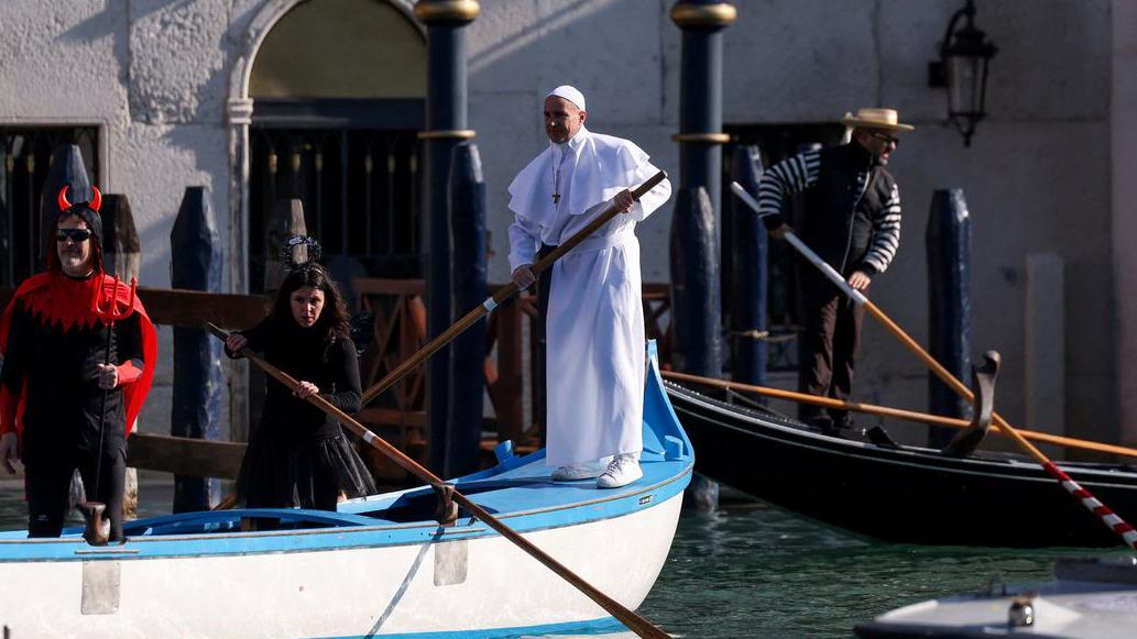 Muškarac obučen kao papa stoji na čamcu i u jednoj ruci drži veslo