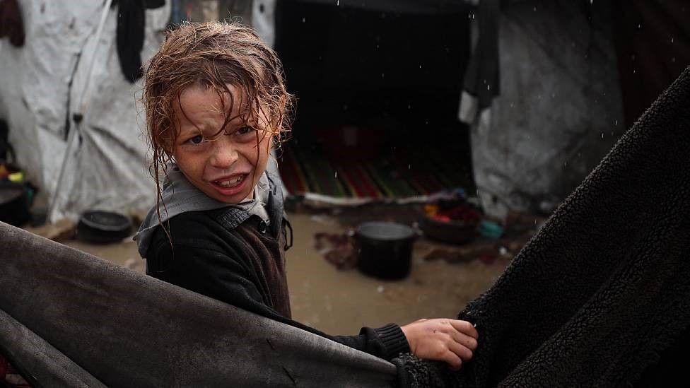 Un niño mira hacia cámara con el pelo y la ropa mojados, por detrás se ve un charco de agua a la entrada de una carpa dañada por la lluvia