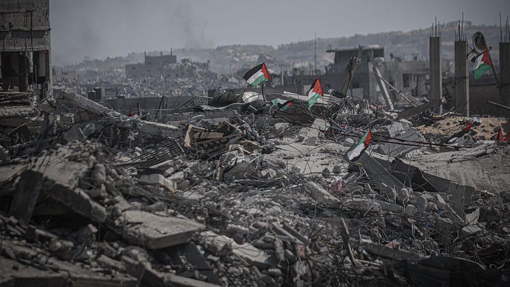 Pilhas de escombros na Faixa de Gaza, sobre as quais se veem bandeiras palestinas. Ao fundo, o terreno est&aacute; totalmente destru&iacute;do e coberto de escombros entre ru&iacute;nas de edif&iacute;cios