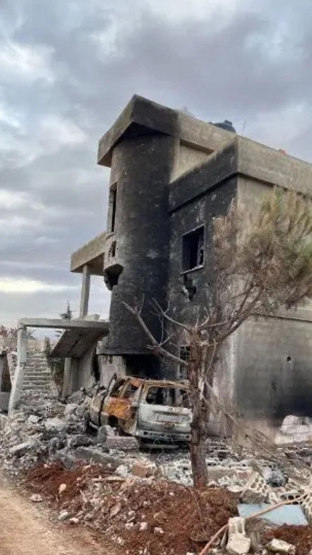 Un edificio de piedra de tres plantas, ennegrecido y derruido, la casa familiar de Qassem, en Líbano, tras los bombardeos, yace en ruinas, con los restos calcinados de un coche y un árbol entre los escombros del exterior, bajo un cielo nublado.