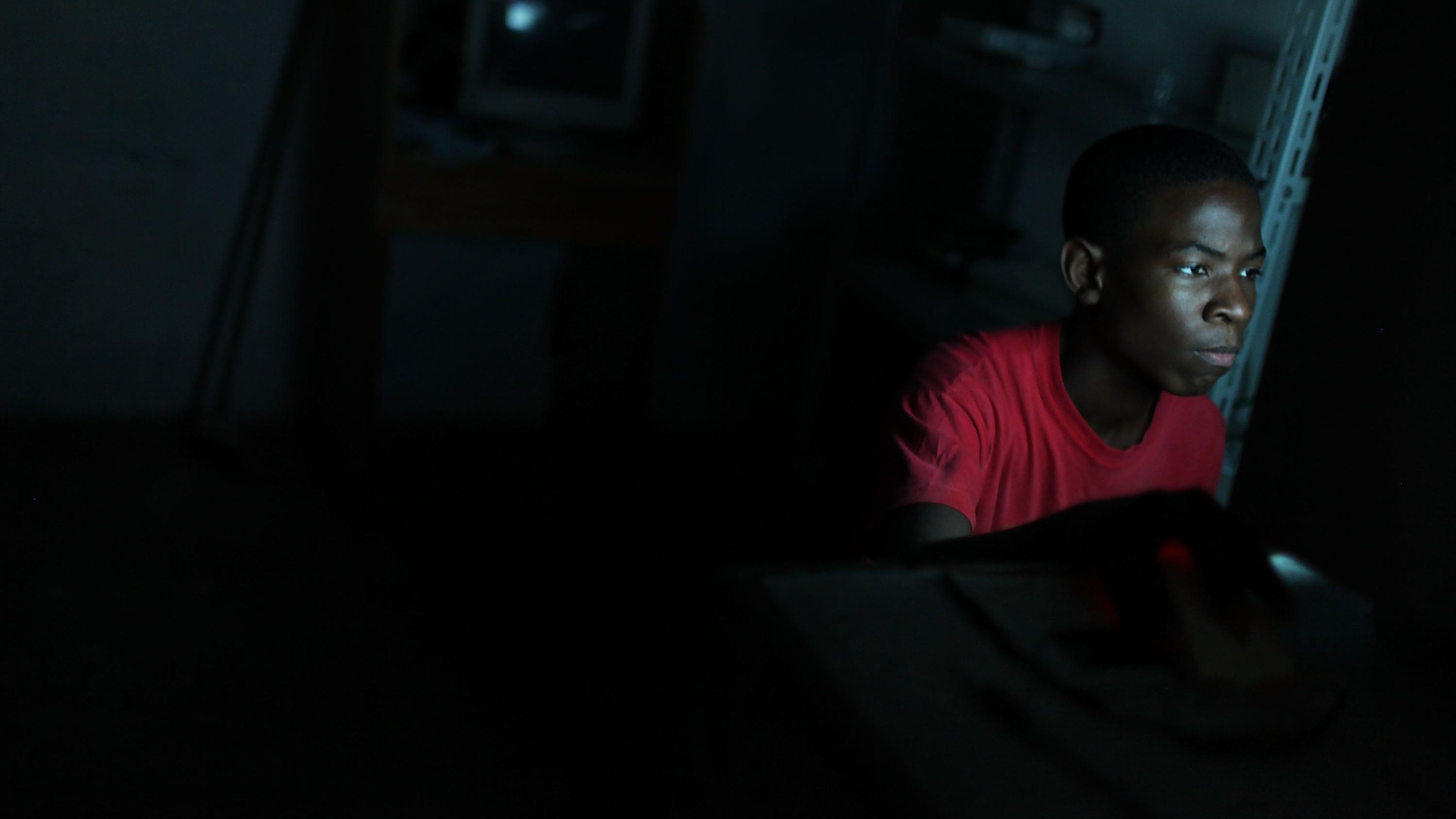 Um jovem aparece sentado diante de uma tela em um ambiente escuro, iluminado principalmente pela luz azulada do monitor que destaca seu rosto concentrado. Vestindo uma camiseta vermelha, ele olha fixamente para a tela enquanto utiliza um computador, cercado por um espa&ccedil;o pouco iluminado e com poucos detalhes vis&iacute;veis ao fundo.