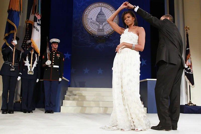 Michelle Obama usa um vestido branco. Ela está com Obama, que dá as mãos a ela em um movimento de dança. 