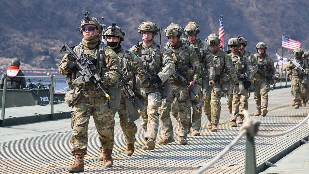 Un contingente militar estadounidense desfila con sus armas. Atrás se ve flameando la bandera norteamericana. 