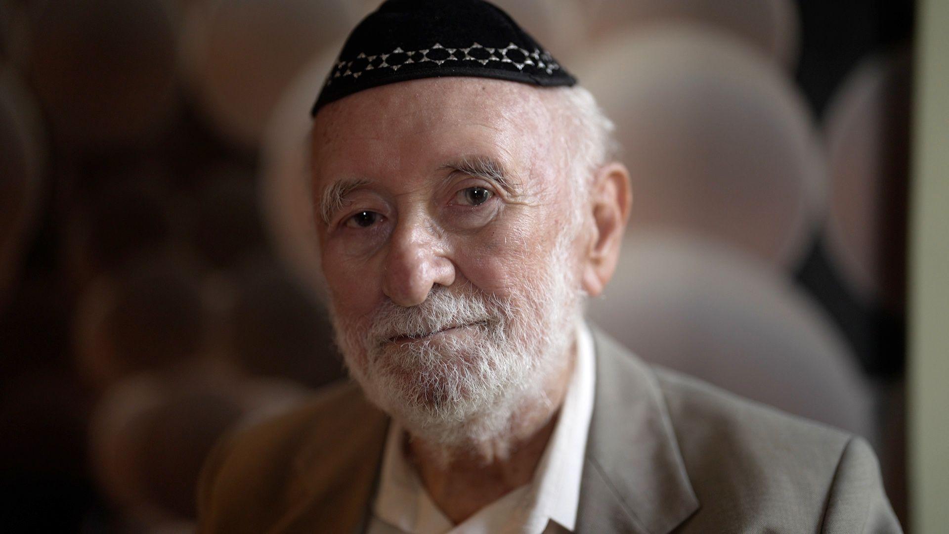 Homem idoso com quipá na cabeça, barba branca e terno.
