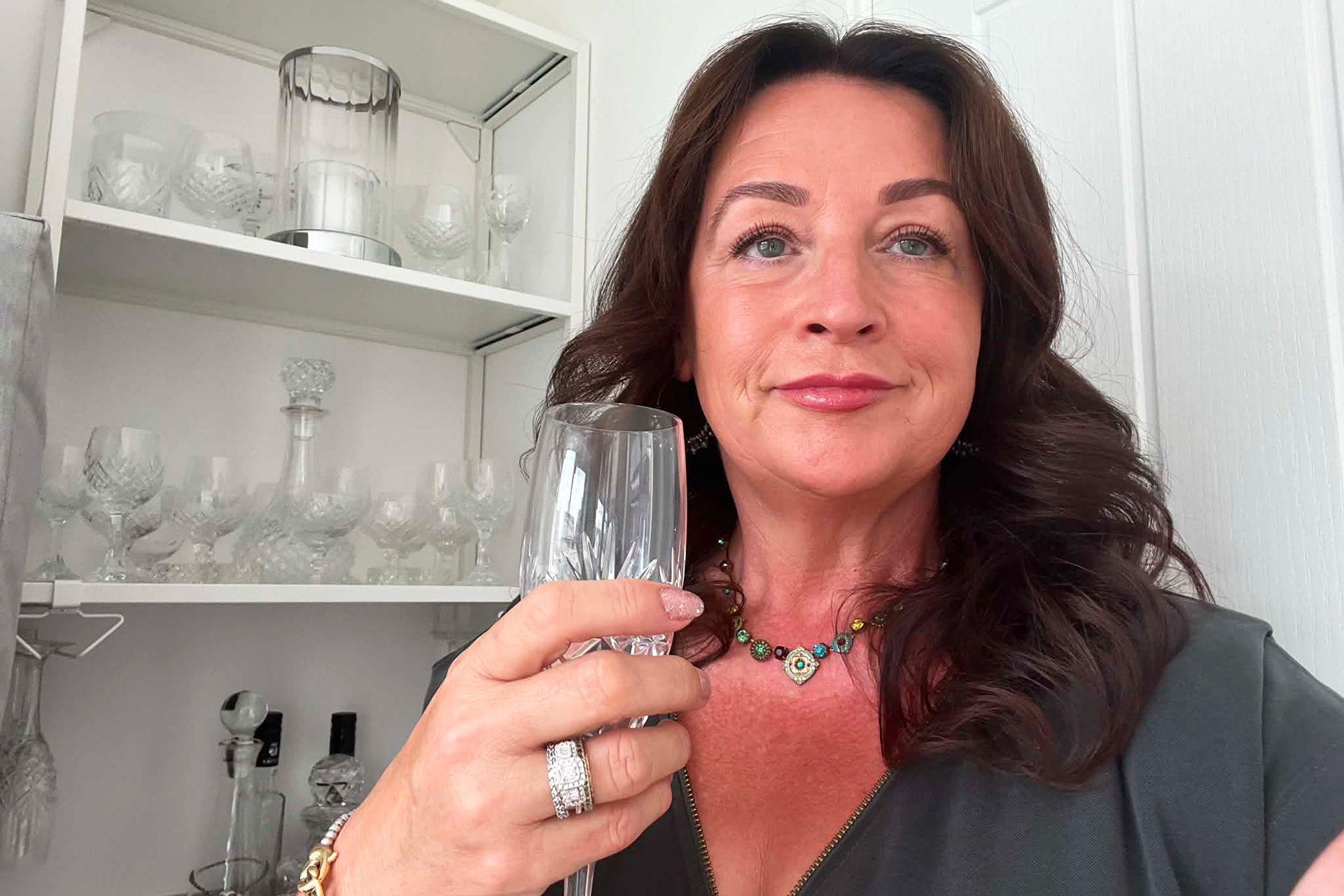 Mulher com longo cabelo escuro e expressão pensativa, segura uma taça de champanhe, com um grande anel visível no dedo anular da mão que segura a taça