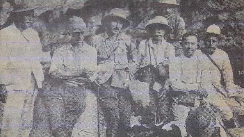 Siete hombres en trajes de exploradores y sombreros de safari posan frente a la cámara recostados sobre una roca