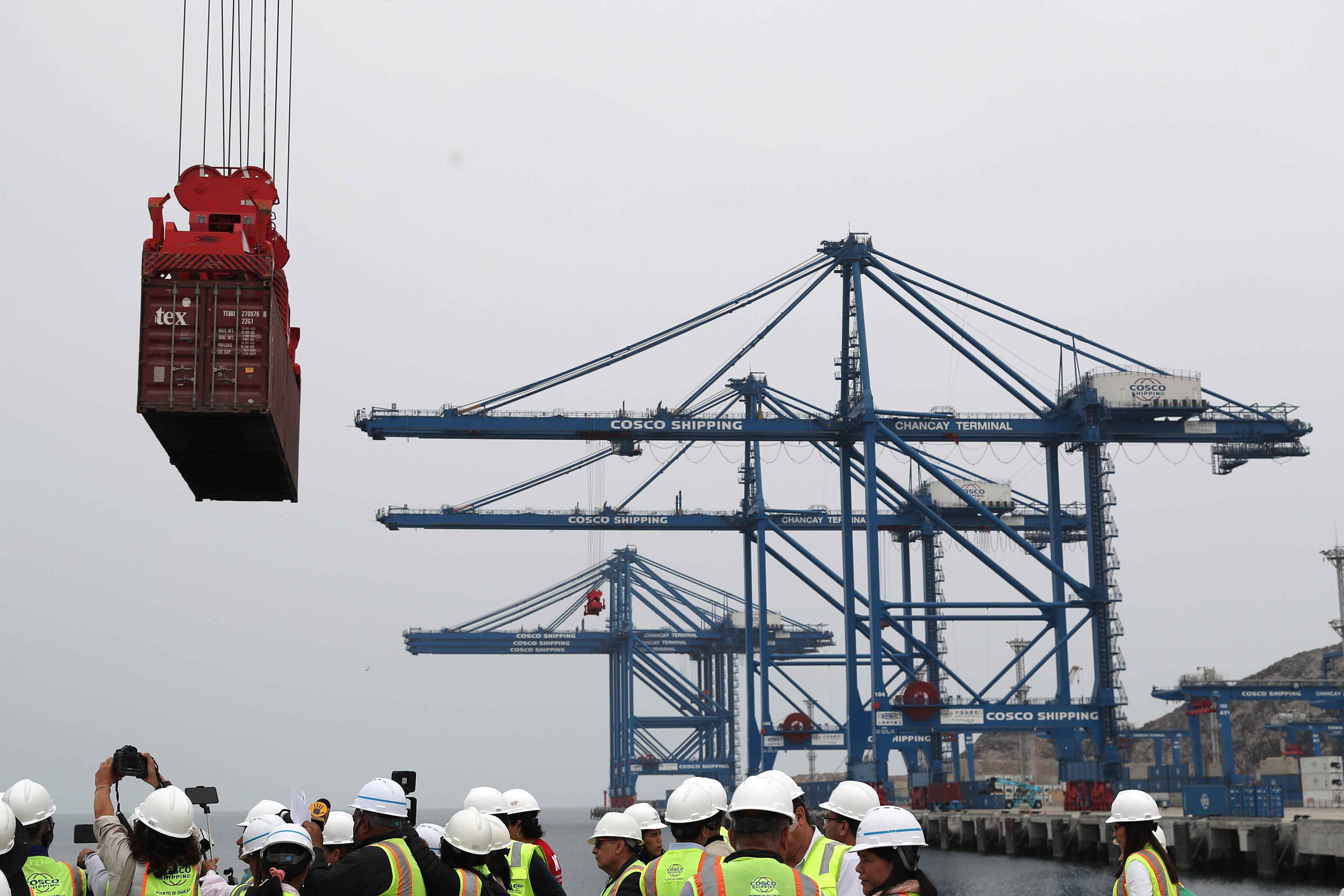 Grúas transportan un contenedor en el gran puerto de Chancay. Debajo, en el suelo, un grupo de personas con cascos y chalecos reflectantes. 