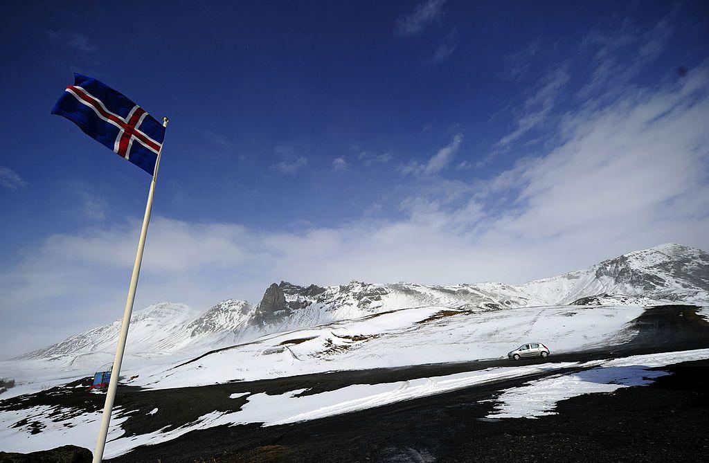 Una bandera de Islandia ondea en lo alto de un mástil en la ladera de una montaña nevada. 