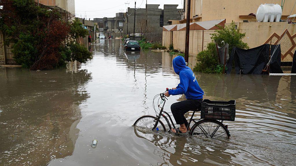 一名身穿藍色連帽衫的年輕男子在摩蘇爾一條被水淹沒的住宅街道上騎單車，遠處可見汽車及灌木叢