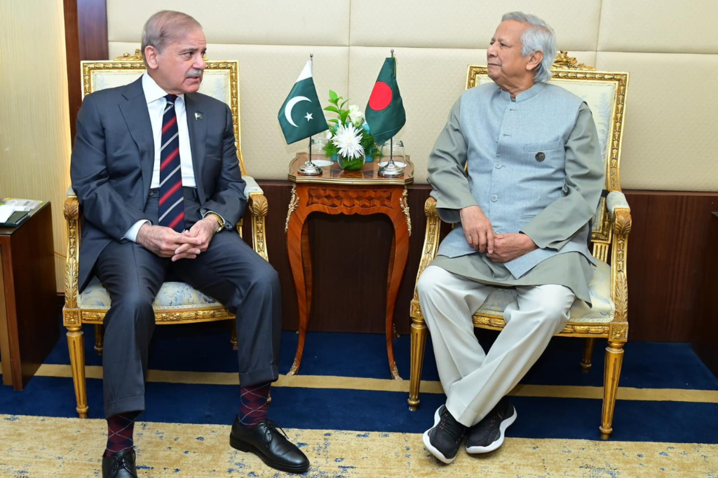 O primeiro-ministro do Paquist&atilde;o, Shehbaz Sharif, aparece sentado ao lado de Muhammad Yunus, chefe do governo interino de Bangladesh. As bandeiras dos dois pa&iacute;ses e algumas flores brancas est&atilde;o sobre uma mesa ornamentada entre os dois