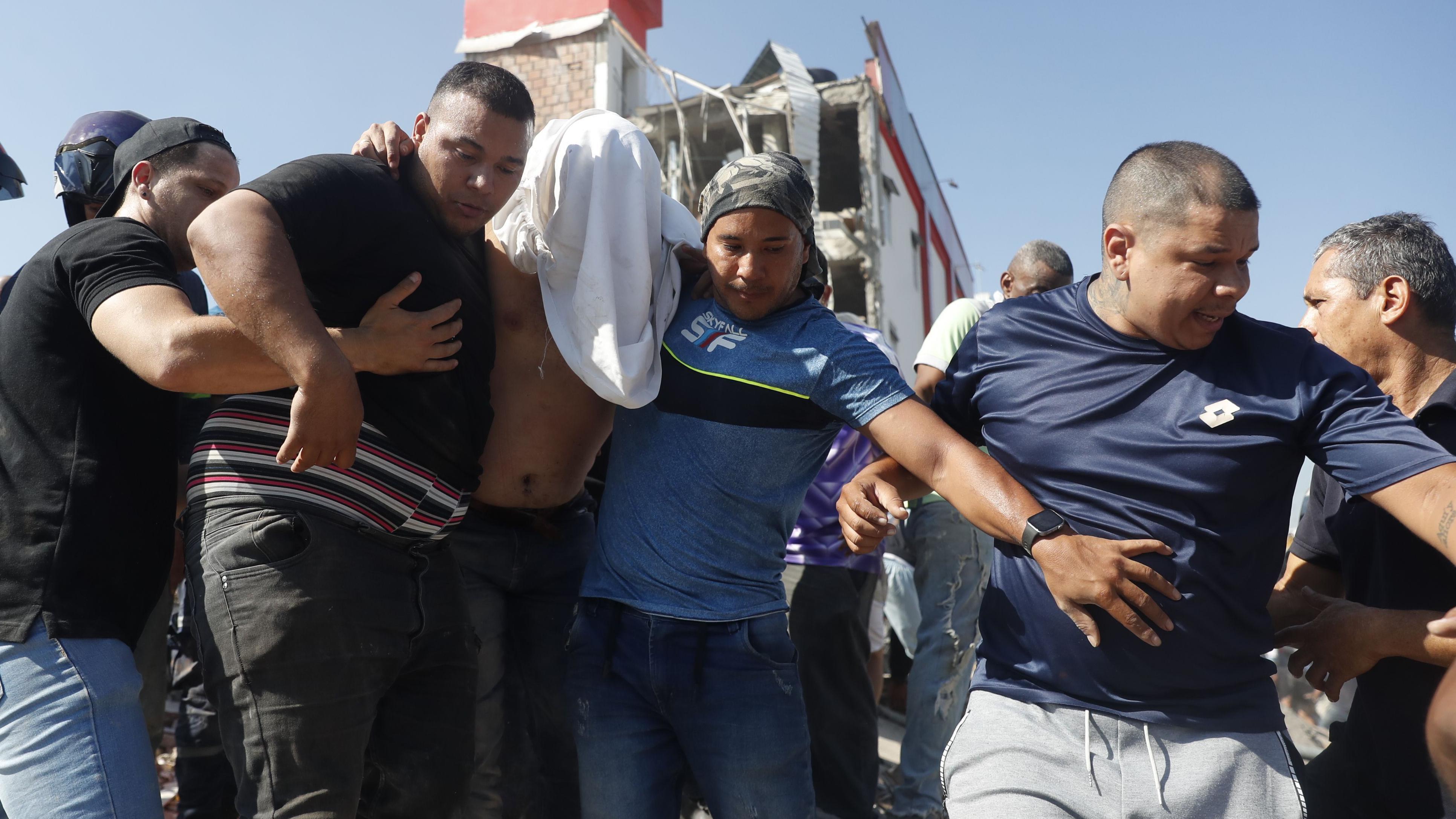 Un hombre, con su camiseta blanca cubriéndole la cara, está siendo ayudado por otros dos hombres a evacuar un edificio colapsado. 