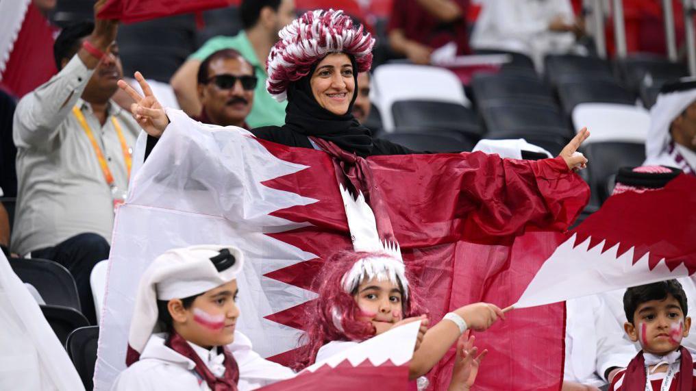 سيدة ترتدي ألوان منتخب قطر التقليدية وامامها أطفال يلوحون بأعلام قطر في مونديال 2022.