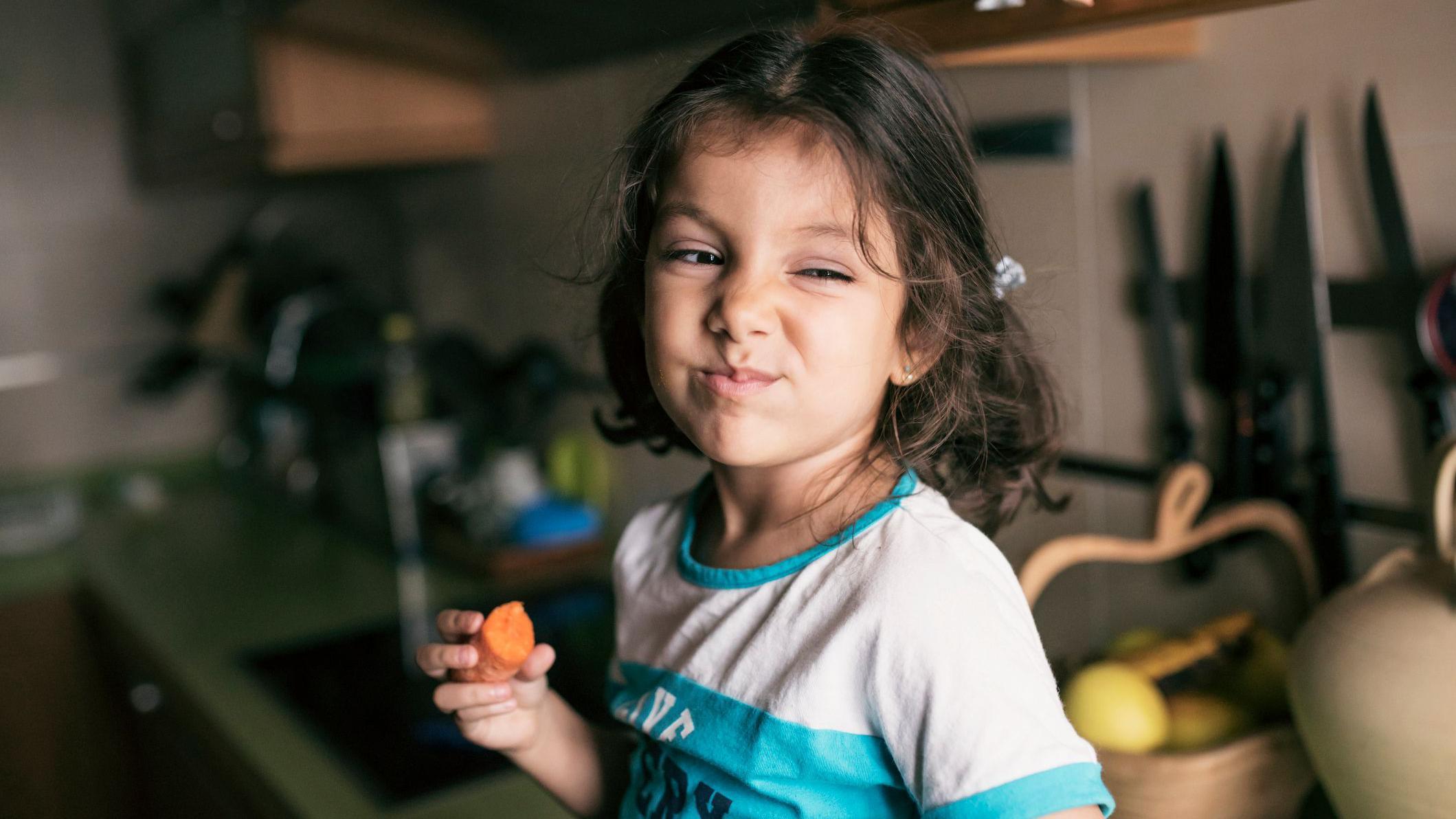 Una niña de 4 años comiendo zanahoria en la cocina, sentada en la encimera.