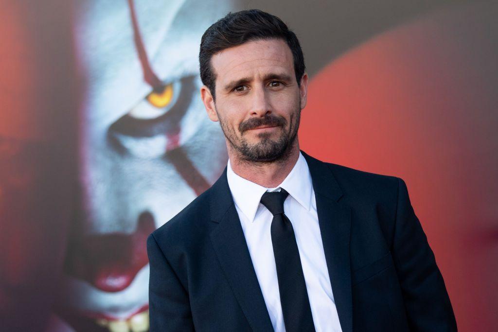 James Ransone de terno preto, camisa branca e gravata preta posa diante de um pôster do filme “It - Capítulo Dois”, com a imagem do palhaço Pennywise ao fundo, durante evento de estreia. 