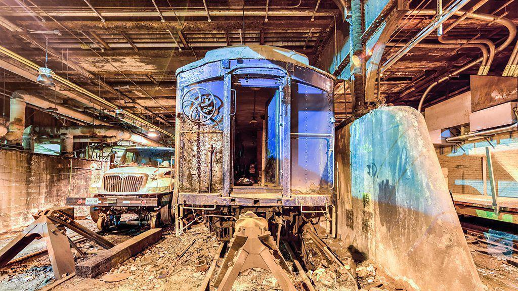 Un viejo vagón de tren que luce oxidado sobre unas vigas, en un túnel sucio lleno de escombros en el suelo. La parte delantera de un camión se ve en el fondo.