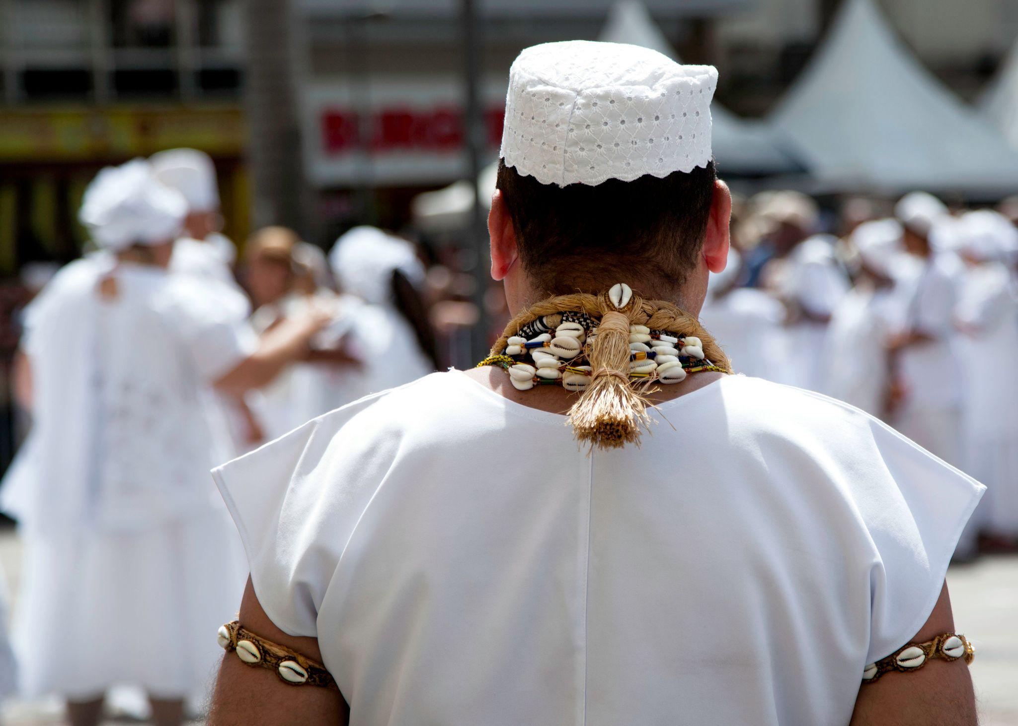 Homem de costas com adorno na cabeça e roupa branca; outras pessoas de branco aparecem ao fundo