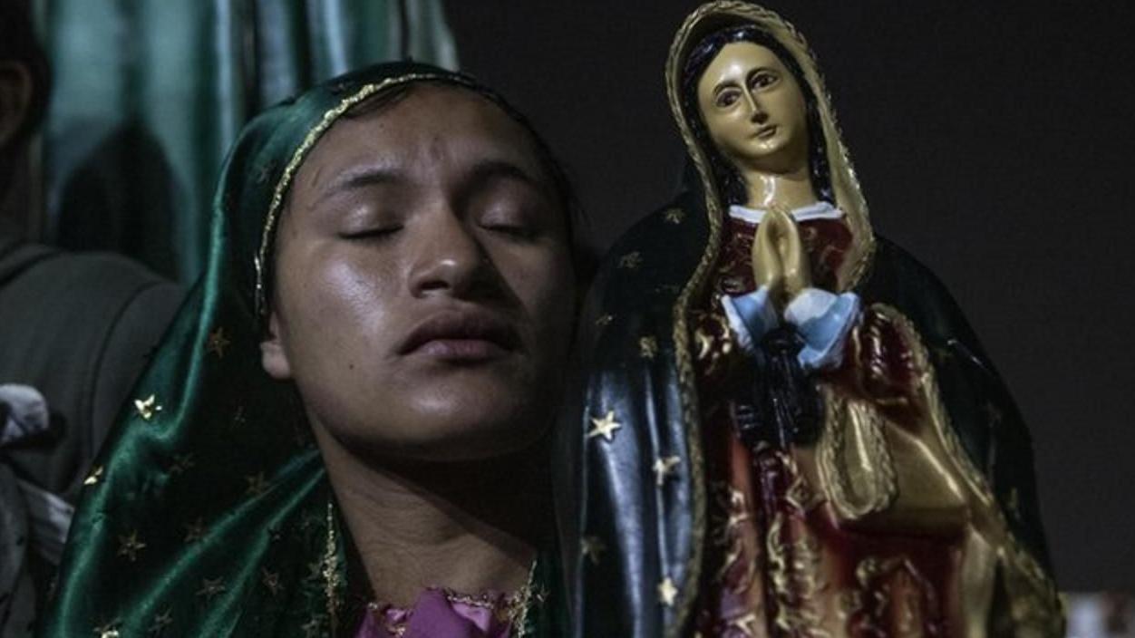 Una mujer devota, con un velo verde con bordes dorados y los ojos cerrados, ora frente a una estatuilla de la Virgen de Guadalupe