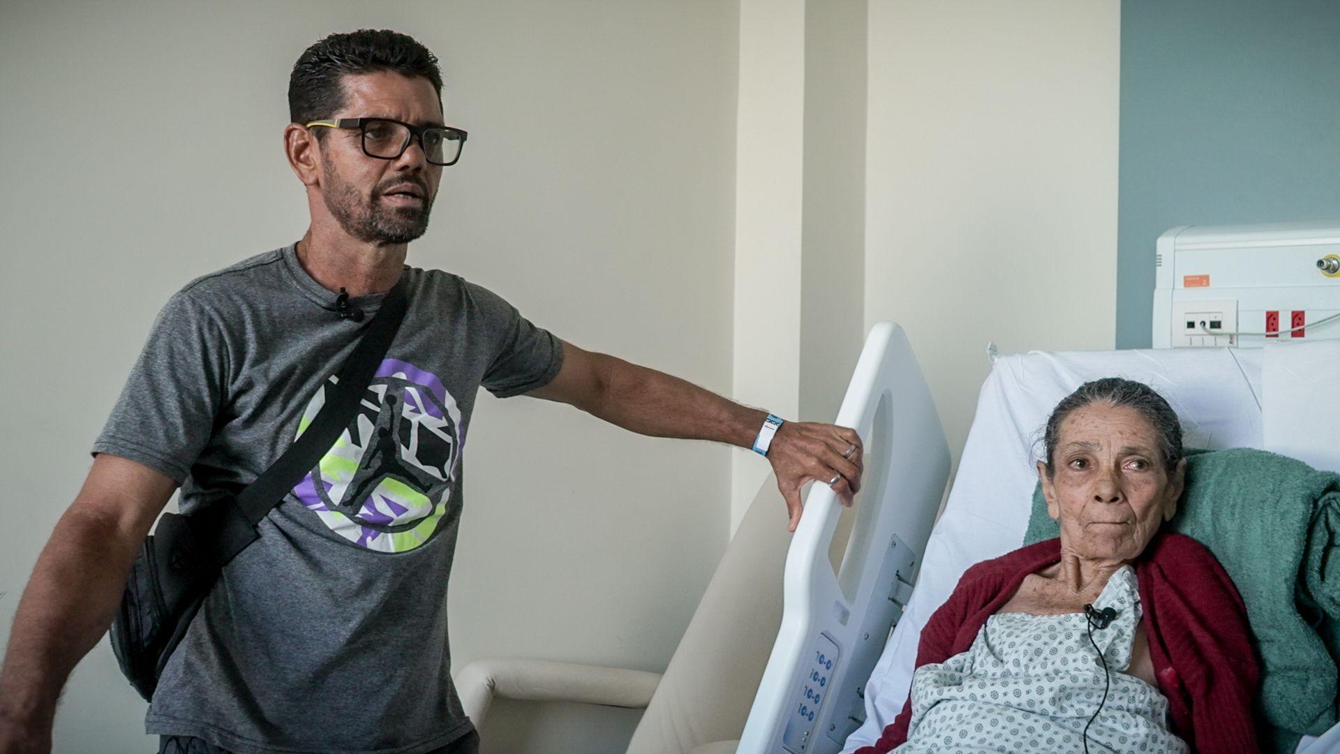 Foto de uma senhora em uma cama de hospital e, ao lado, de pé, um homem apoiado na cama.