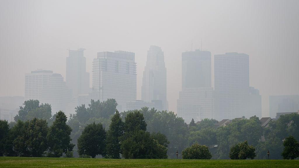 El horizonte de Minneapolis, Minnesota, está borroso por el humo, con el césped verde del parque y los árboles en primer plano.