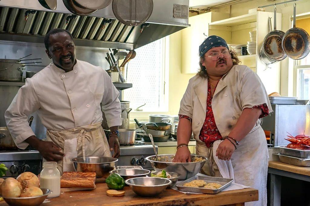 Michael Jibrin, en su papel de Tyrone y Stavros Halkias, interpretando a Dimitri. Están en medio de una cocina con ingredientes sobre la mesa.