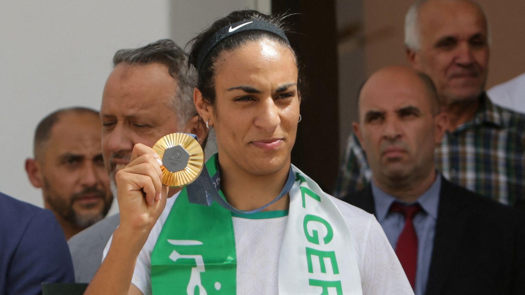 Imane Khelif sostiene una medalla. Hay varios hombres detrás de ella.