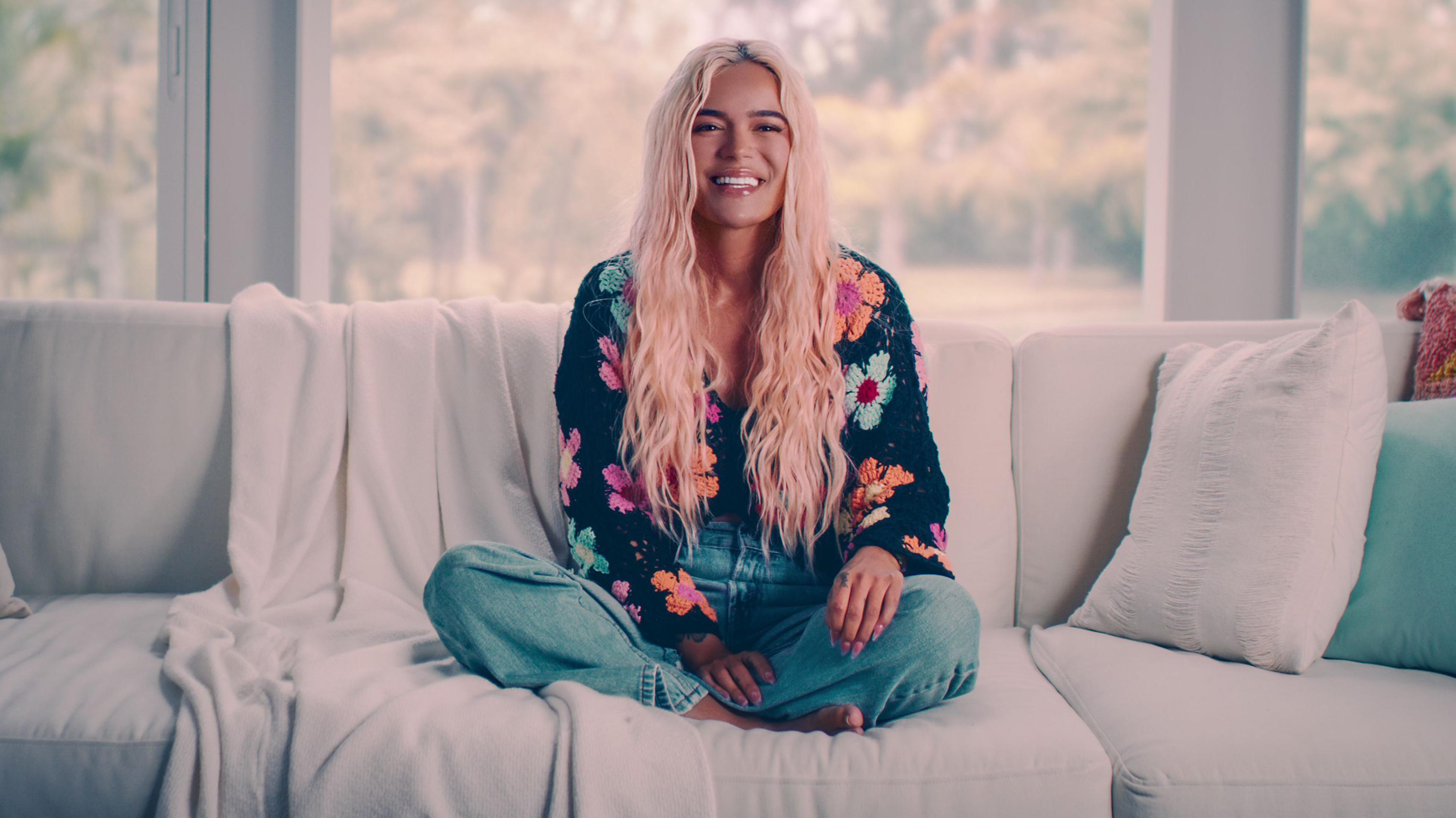 karol G sonríe, sentada en un sofá