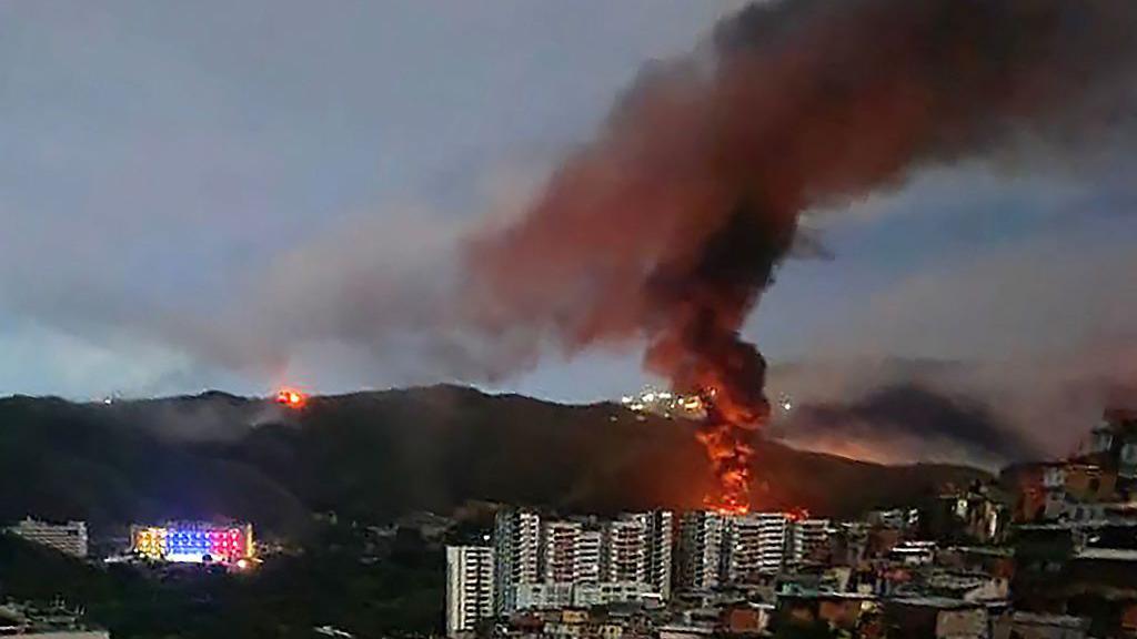Un incendio en Fuerte Tiuna, el complejo militar más grande de Venezuela, se observa a la distancia tras una serie de explosiones en Caracas. Se ve una columna de humo elevándose hacia un cielo oscurecido, detrás de varios edificios altos y con una montaña al fondo.