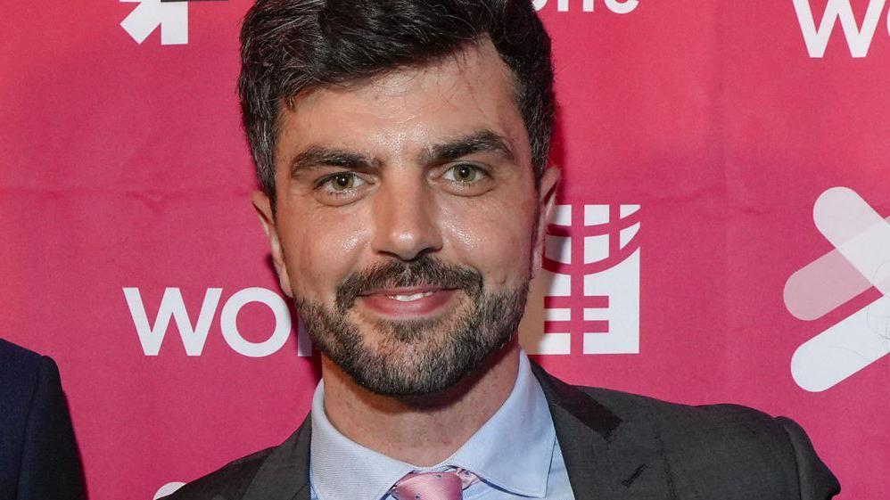 Will Adolphy, un hombre con barba bien cuidada y pelo corto, vestido con una chaqueta oscura, camisa azul y corbata rosada, sonríe directamente a cámara