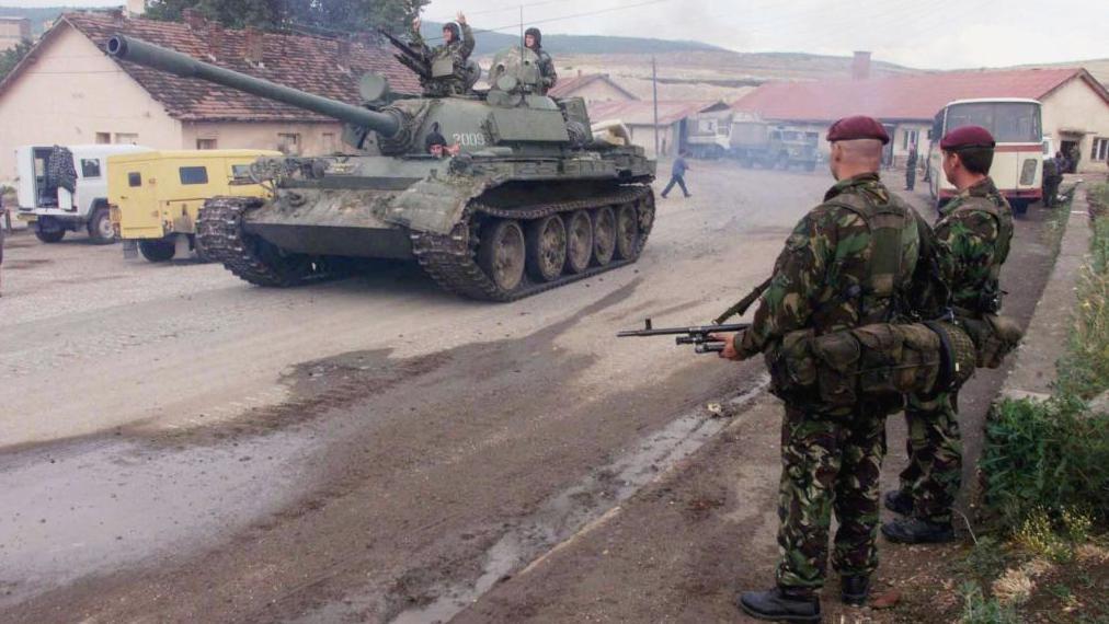 A Serbian tank and its crew drive past standing British soldiers in Kosovo