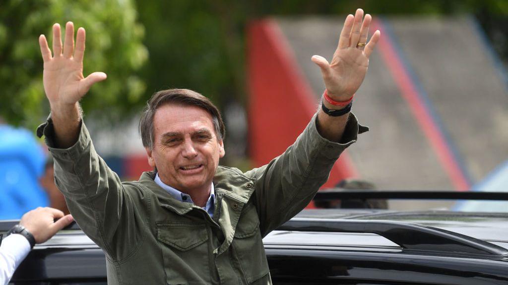 Imagem de Jair Bolsonaro na rua, com as m&atilde;os para cima, cumprimentando o p&uacute;blico. 