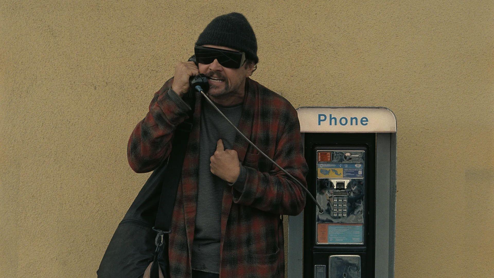 Leonardo Di Caprio vistiendo un gorro de lana, lentes oscuros y una bata hablando en un teléfono público