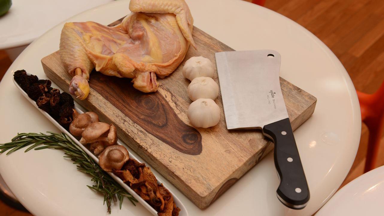 A butcher's knife with a black handle placed next to 3 garlic bulbs and a uncooked chicken on top of a cutting board