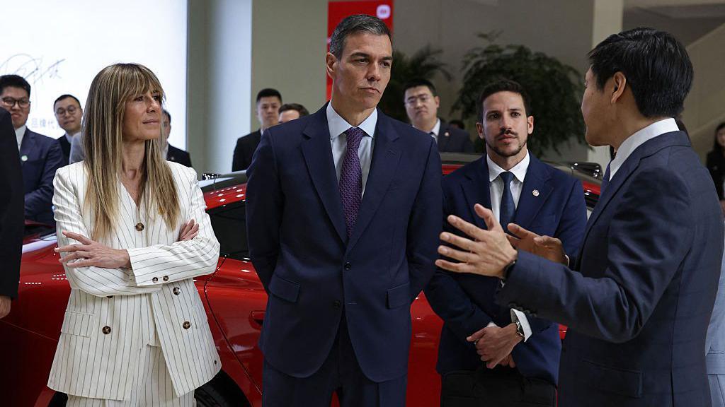 Pedro Sánchez, en medio, vestido con traje y corbata conversa con un funcionario chino (a la derecha) durante un viaje oficial. Begoña está de pie a la izquierda con los brazos cruzados. Es una mujer rubia, delgada y viste un chaleco color blanco. En el fondo se ven varios hombres también vestidos de traje y un auto rojo. 