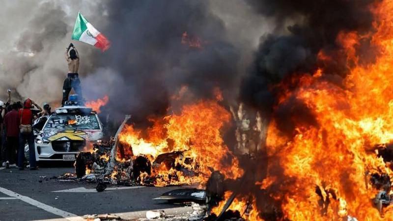 Manifestante no alto de um carro exibe uma bandeira do México, enquanto veículos são incendiados em uma rua de Los Angeles, em 8/6/2025