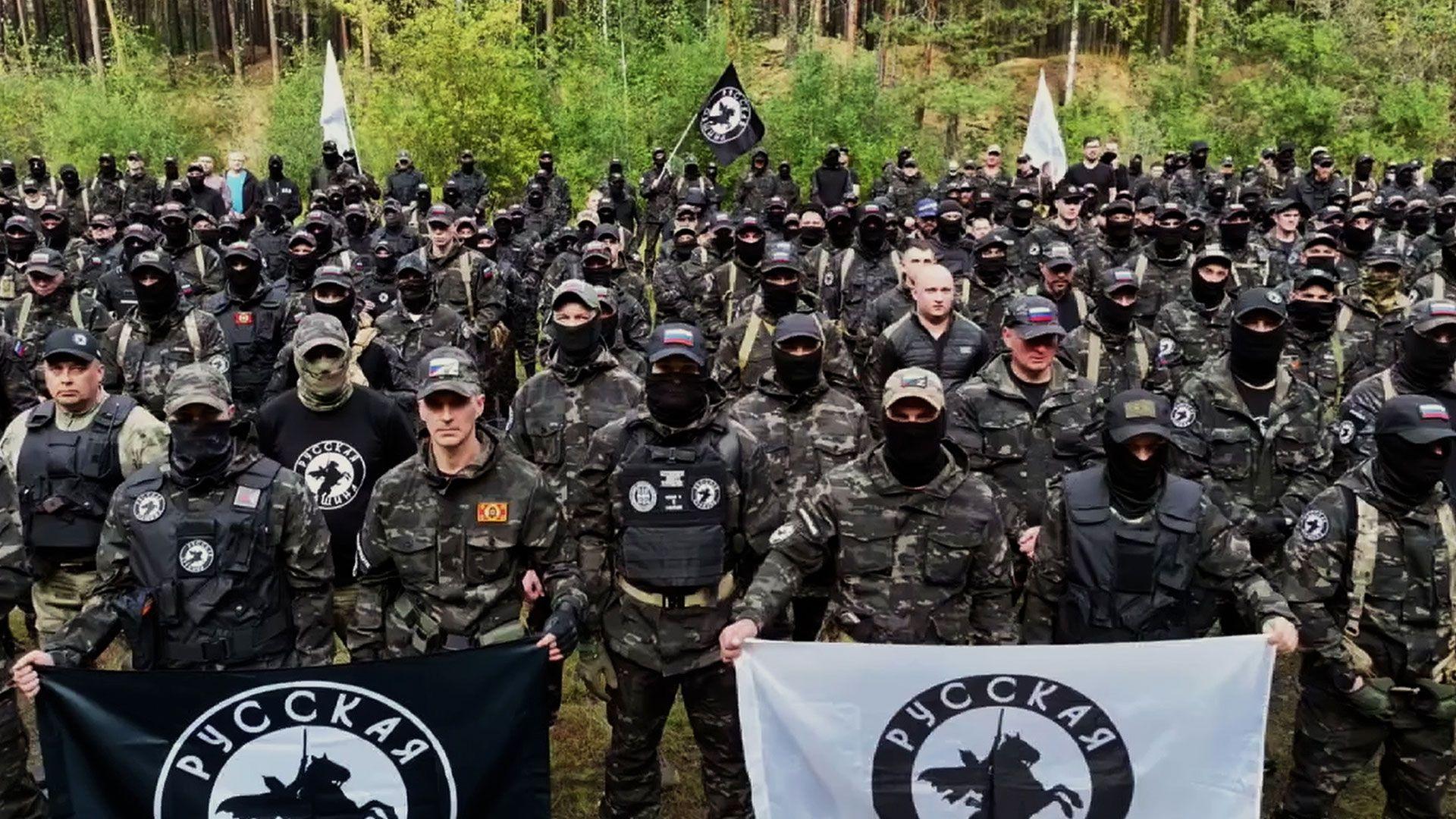 Dezenas de homens usando roupas militares e segurando bandeiras pretas e brancas com a imagem de um cavaleiro montado posam diante da c&acirc;mera, com uma &aacute;rea de floresta ao fundo