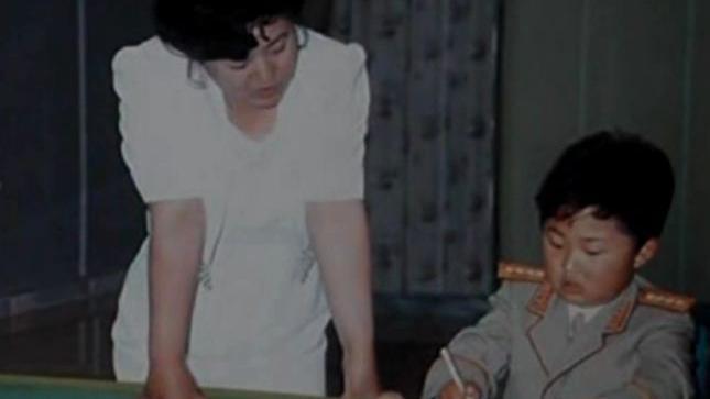 Ko Yong-hui, em um vestido branco, &agrave; esquerda. Ao lado dela, sentado, est&aacute; Kim Jong-un, ainda crian&ccedil;a, vestindo uniforme militar e escrevendo com uma caneta. 