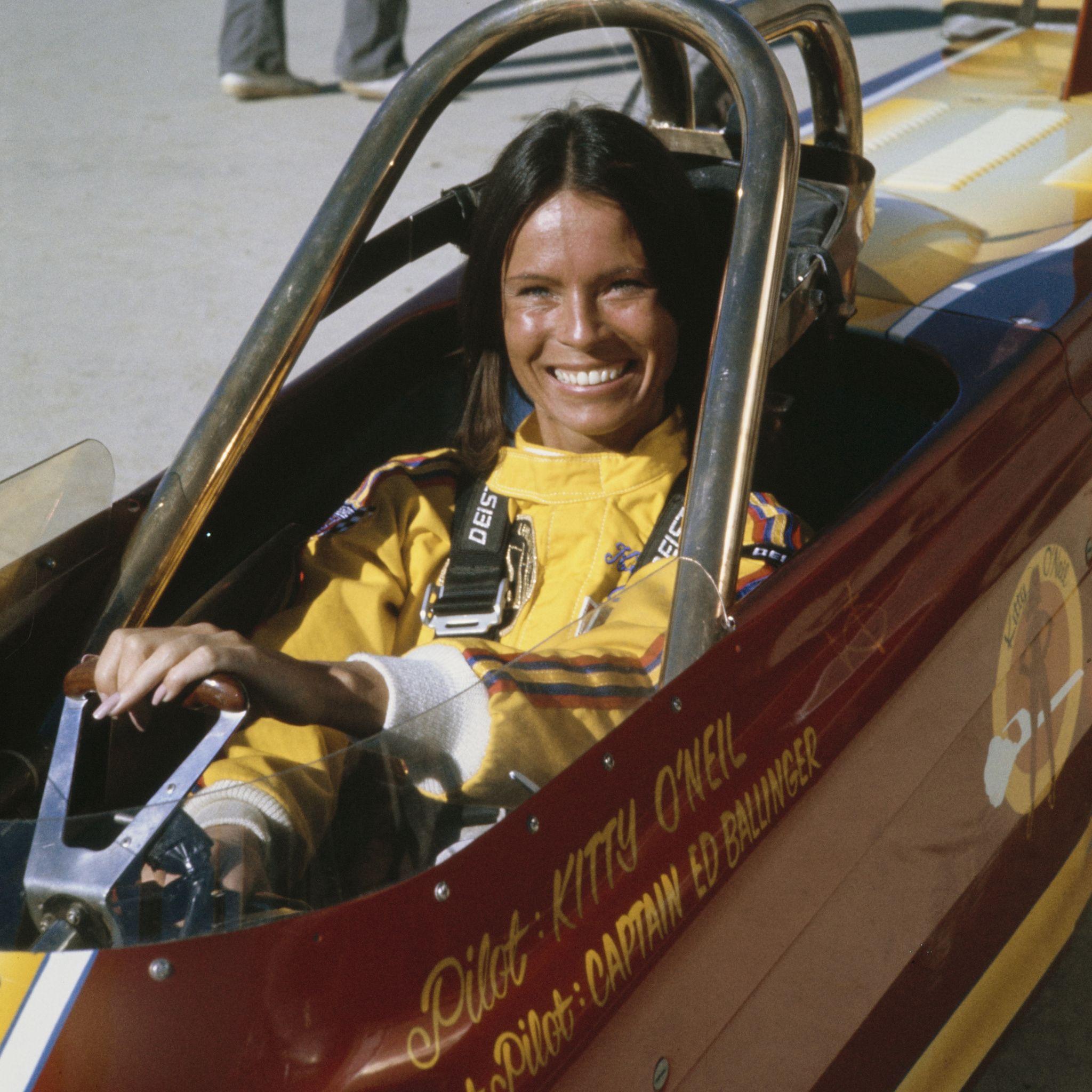基蒂·奧尼爾（Kitty O'Neil）——一位留著烏黑直髮的女性——坐在一輛敞篷車中，身穿黃色連身衣，雙手握著方向盤，對著鏡頭露出微笑（7/7/1977）