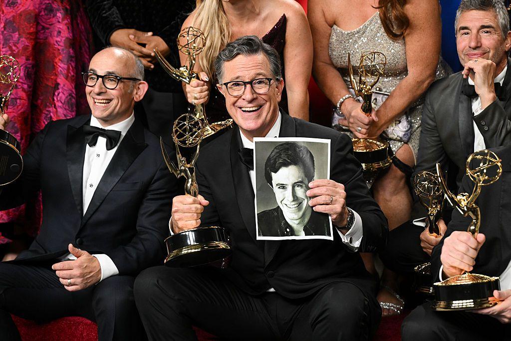 Stephen Colbert mira al frente, vestido de etiqueta sostiene el Emmy en su mano derecha y una foto suya de cuando era joven en la mano izquierda. A su alrededor hay otros ganadores con sus estatuillas en la mano.
