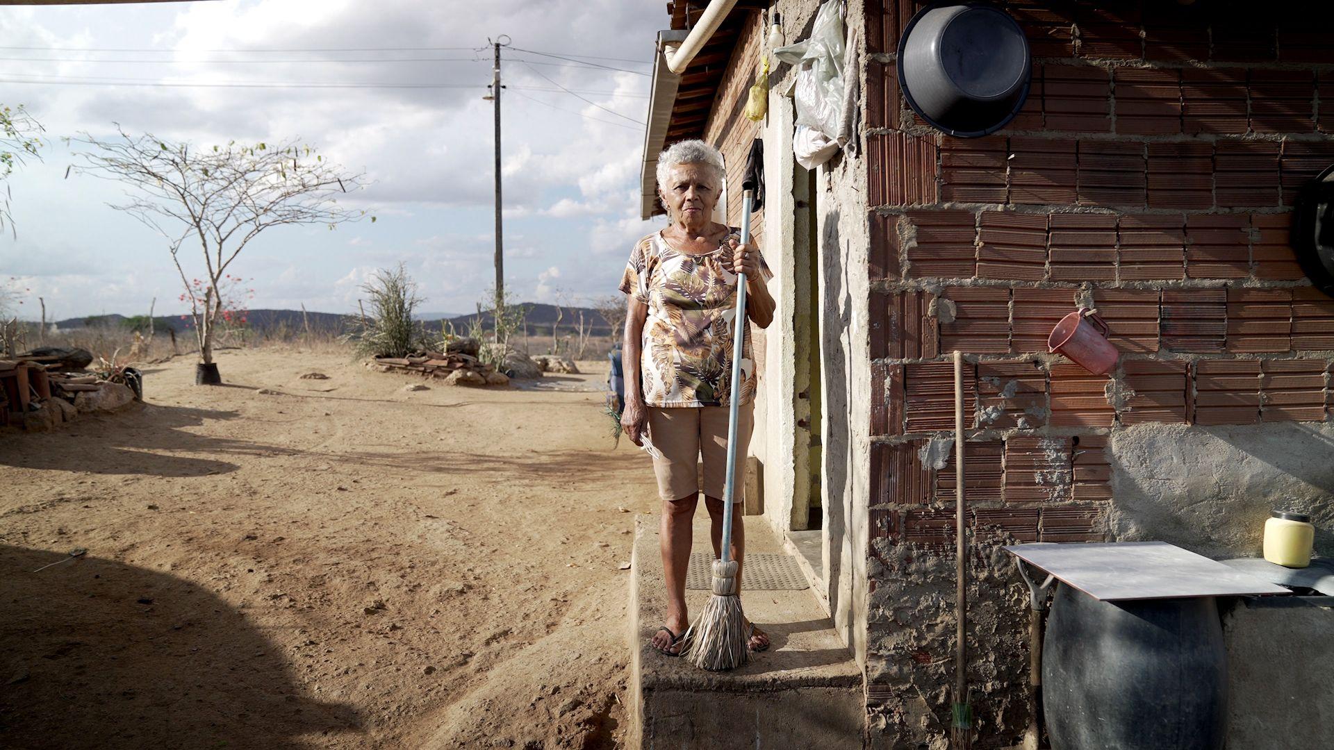Mulher idosa de cabelos brancos com vassoura à porta de casa simples, em paisagem árida