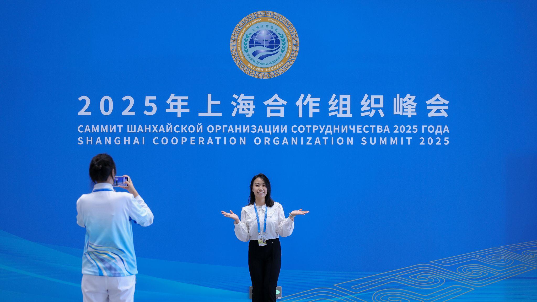 天津上合組織峰會新聞中心內兩位女性工作人員站在有大會標識的淺藍色佈景板前拍照留念（中新社圖片28/8/2025）
