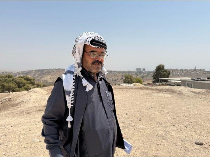أبو عماد يرتدي ثوباً رمادي اللون وعلى رأسه الكوفية الفلسطينية، يقف في منطقة ترابية.