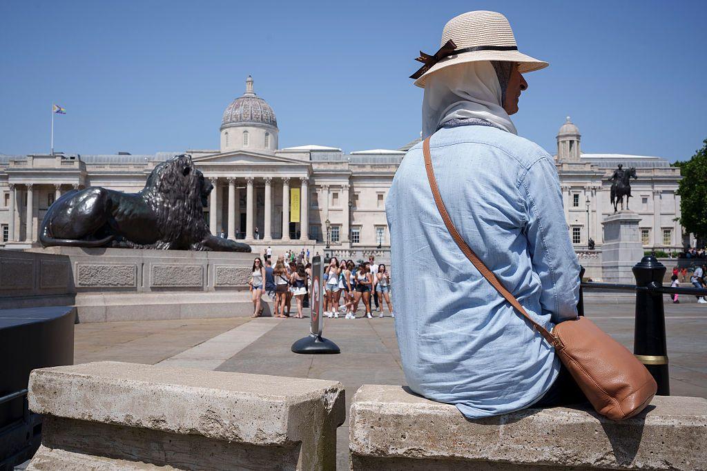 Mujer con camisa delgada y holgada de manga larga, protegida con un pañuelo y un sombrero, está sentada frente a la Galería Nacional de Londres. 
